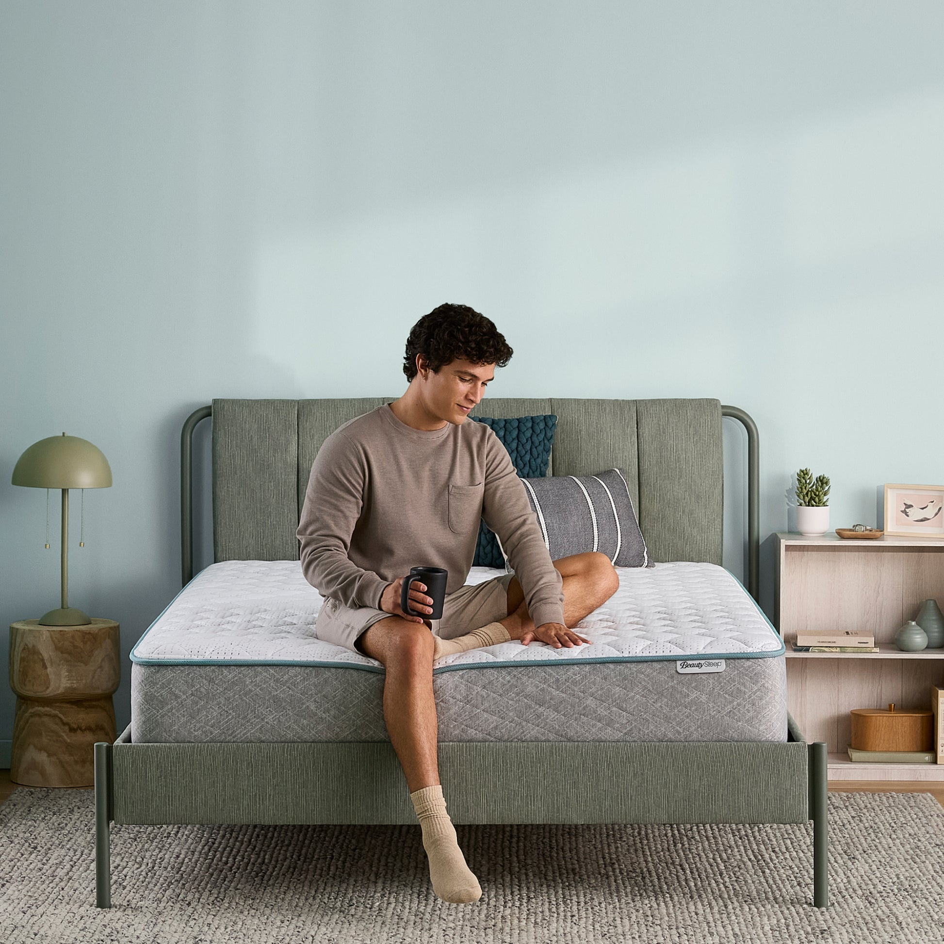 Man sitting on a Beauty Sleep mattress, holding a mug in a bedroom setting, rubbing on the mattress.