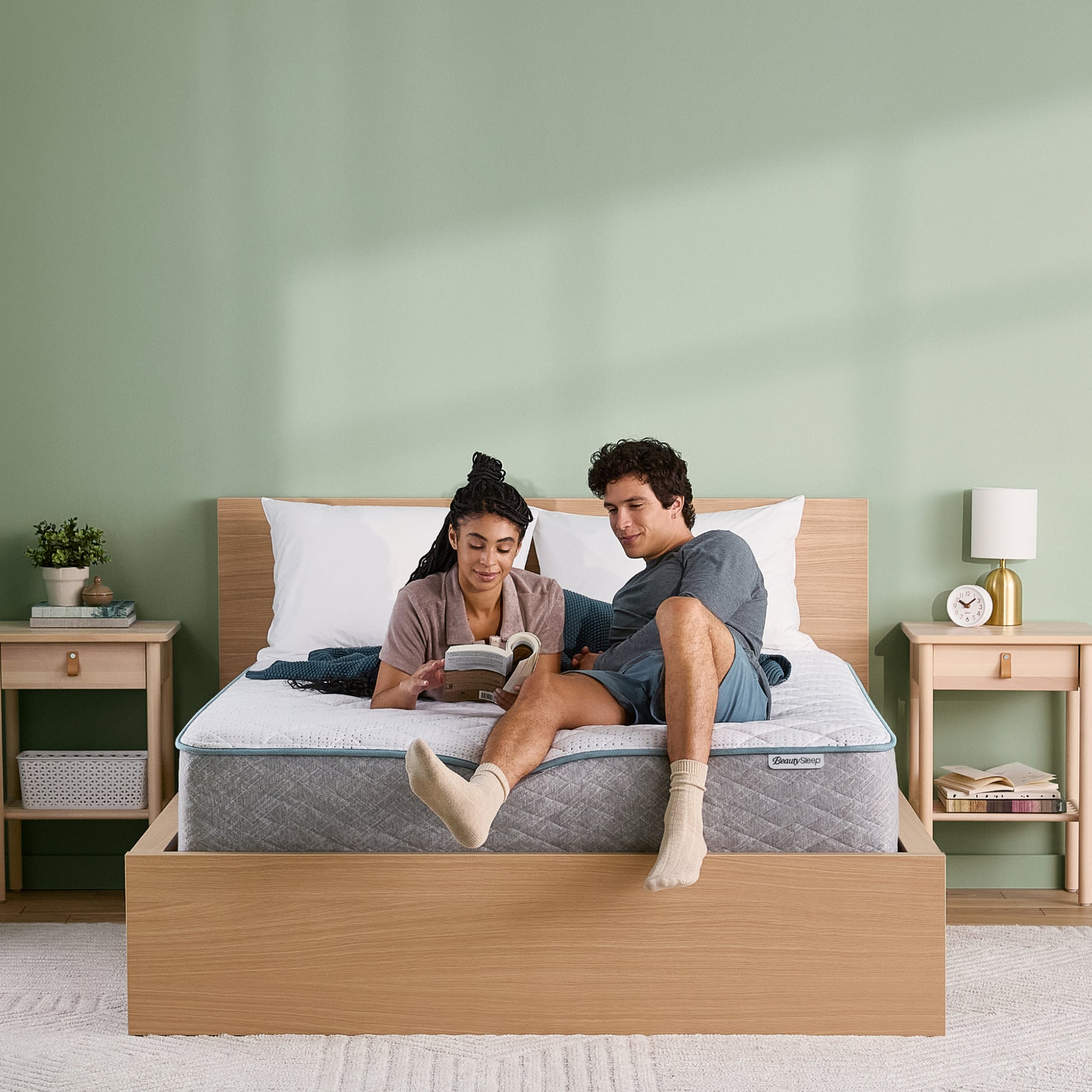 Couple sitting on a Beauty Sleep mattress in a wooden bed frame in a bedroom with a green wall.