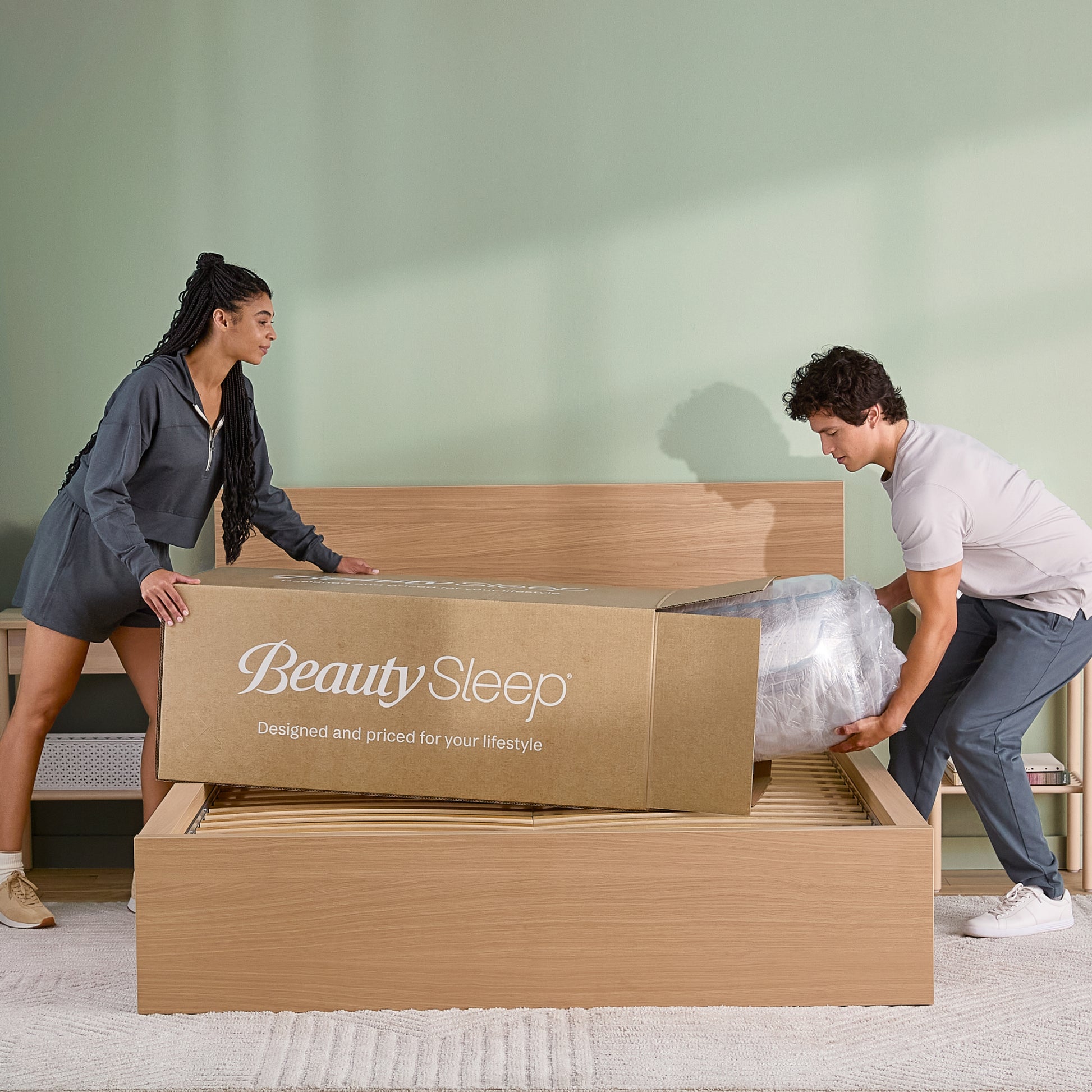Two people unboxing a 'BeautySleep' mattress in a room with a green wall.
