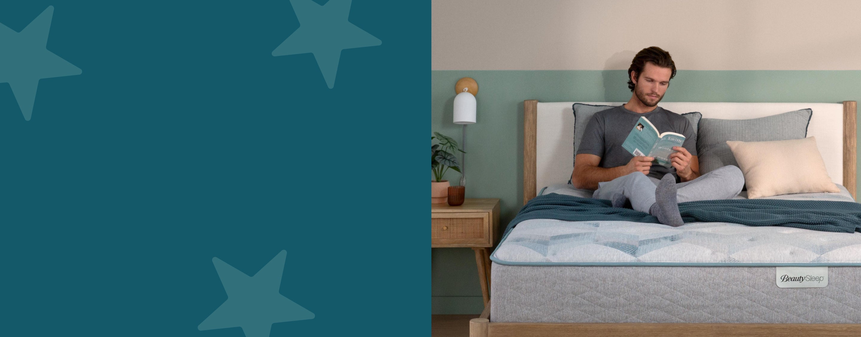 Man sitting on top of a Beauty Sleep mattress reading a book while leaning on 1 of 3 pillows and a blanket on top of the mattress.